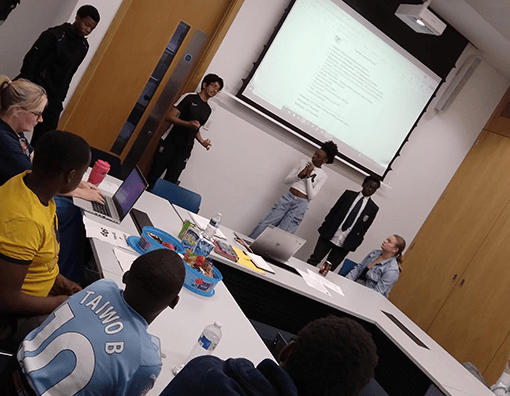 Photograph of a group presentation in a classroom setting with students seated around a U-shaped table and three presenters standing near a projection screen displaying text. The scene includes laptops, notebooks, and water bottles on the table, highlighting an interactive academic or collaborative environment.