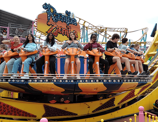 Photograph of a group of young people seated on an amusement park ride featuring bright orange seats and a yellow track structure. The ride platform is decorated with colourful lights, and the riders display a mix of excitement and anticipation.