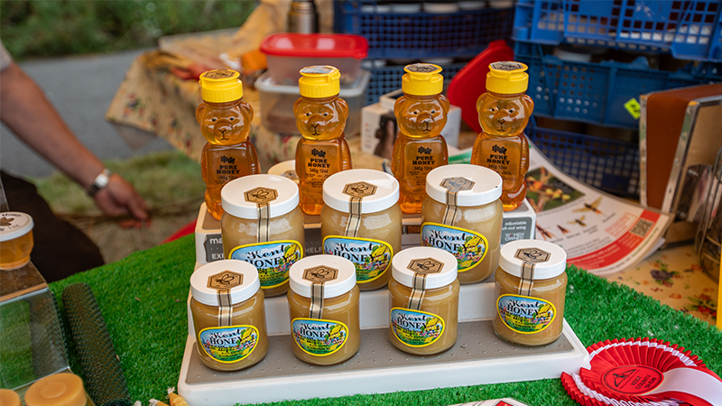 A market stall display of local Kent honey in glass jars and bear-shaped bottles, alongside beeswax candles and a Kent County Show rosette.