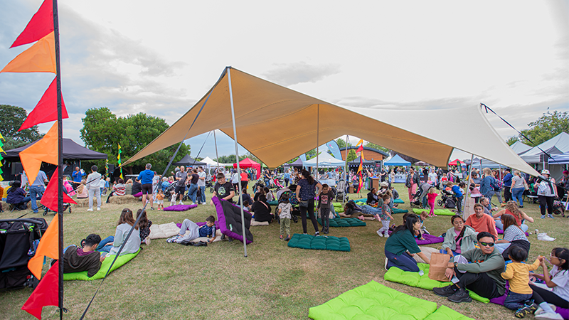 Families relax on colourful cushion mats under a cream stretch tent at a busy outdoor community festival lined with stalls and bunting.