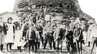  A black and white photograph showing a group of children and adults gathered in front of a large, circular structure. The scene captures early 20th century clothing styles and a communal or educational event.