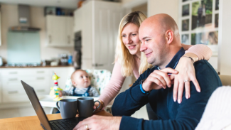 A couple with their baby attend an online fostering information session from their kitchen table