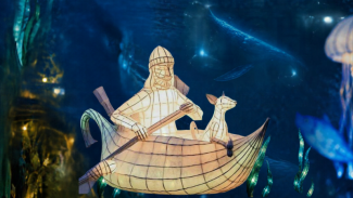 Illuminated paper lantern sculpture of a person rowing a boat with a dog, with the sea and whales swimming in the background. 